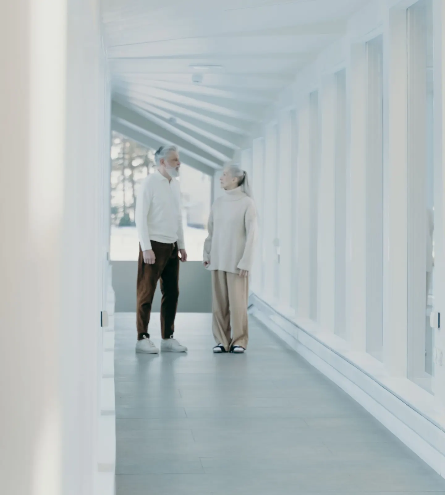 An older couple talking in a bright, white hallway lined with windows.