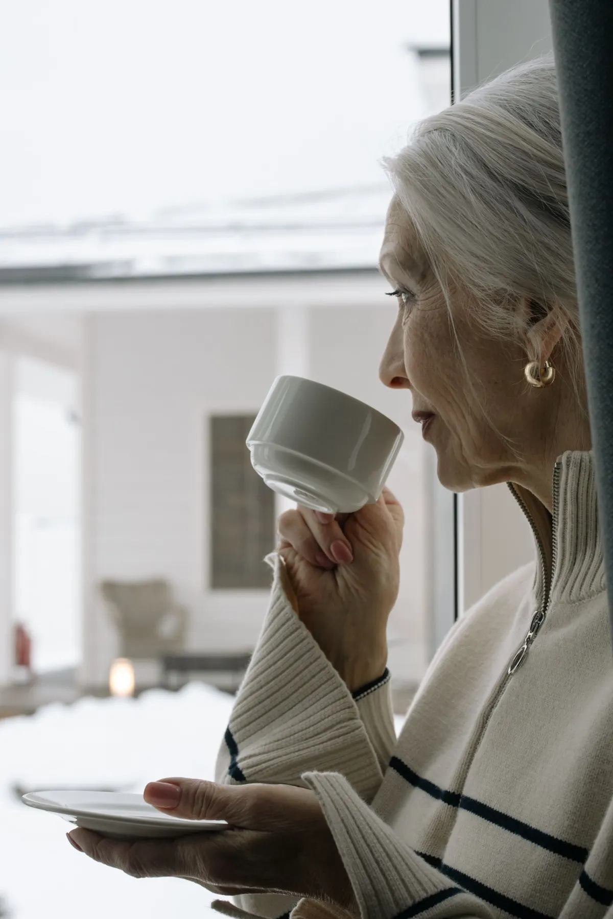 An older woman with a cup of coffee by a snowy window