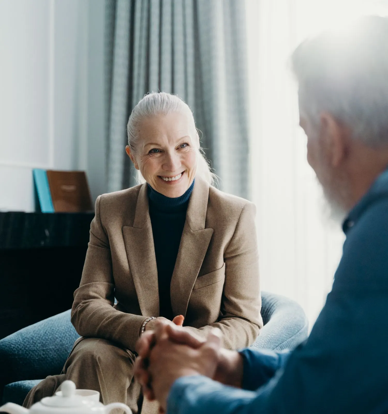 An older woman smiling as she talks with an older man over tea in a bright living room